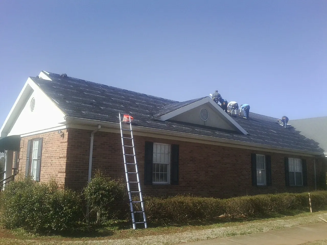 Crew tearing shingles off a brick ranch roof with a ladder set — Metal Roof Repair work by our crew in New Lisbon